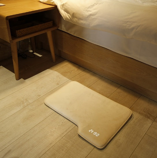 Beige bath mat with digital clock on a wooden floor next to a bed